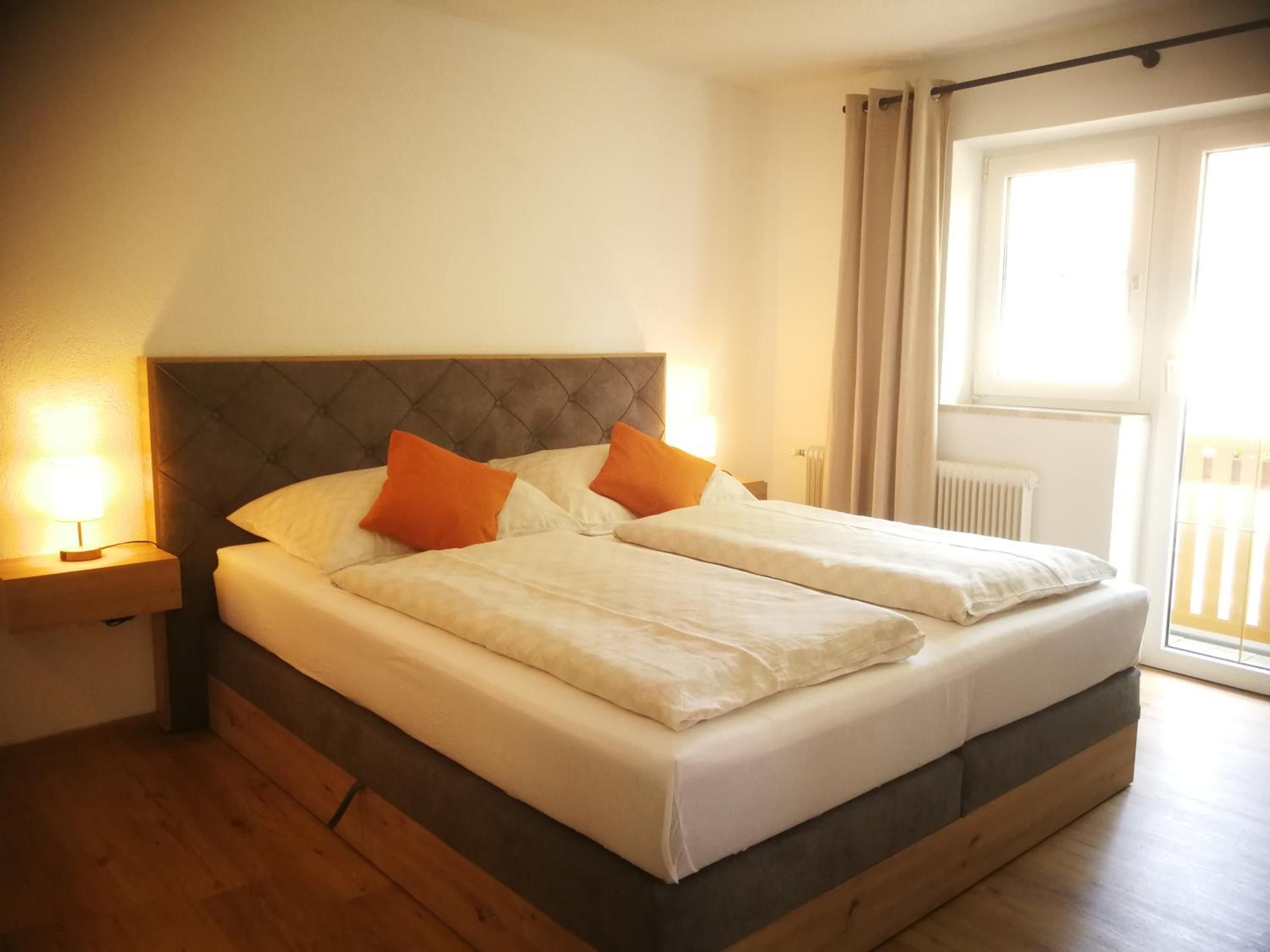 Ein Doppelbett mit Kopfteil, zwei Kissen, ein Nachttisch mit Lampe und ein Fenster mit Vorhängen.