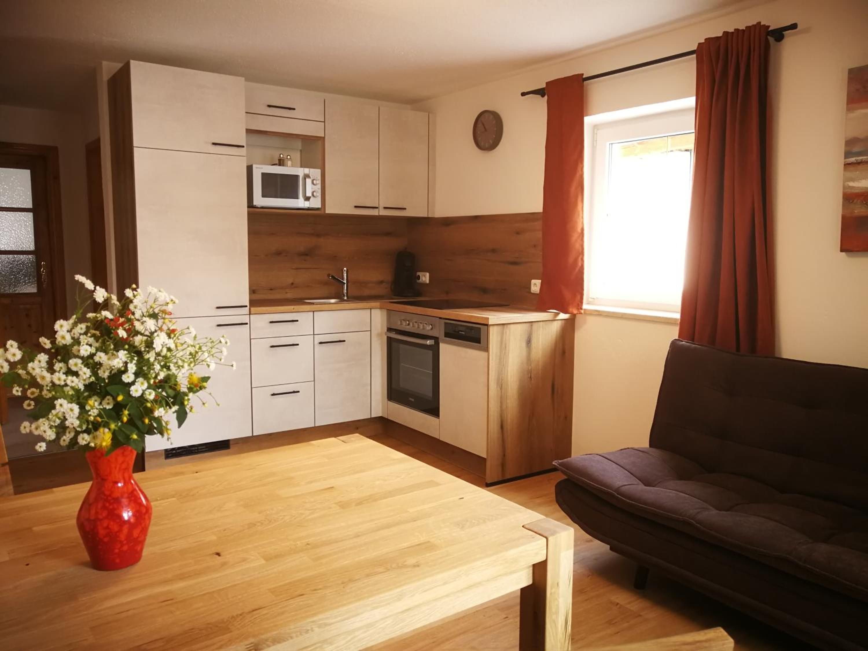 A compact kitchen with a microwave, oven, and sink, a dining table with a vase of flowers, and a dark-colored sofa.