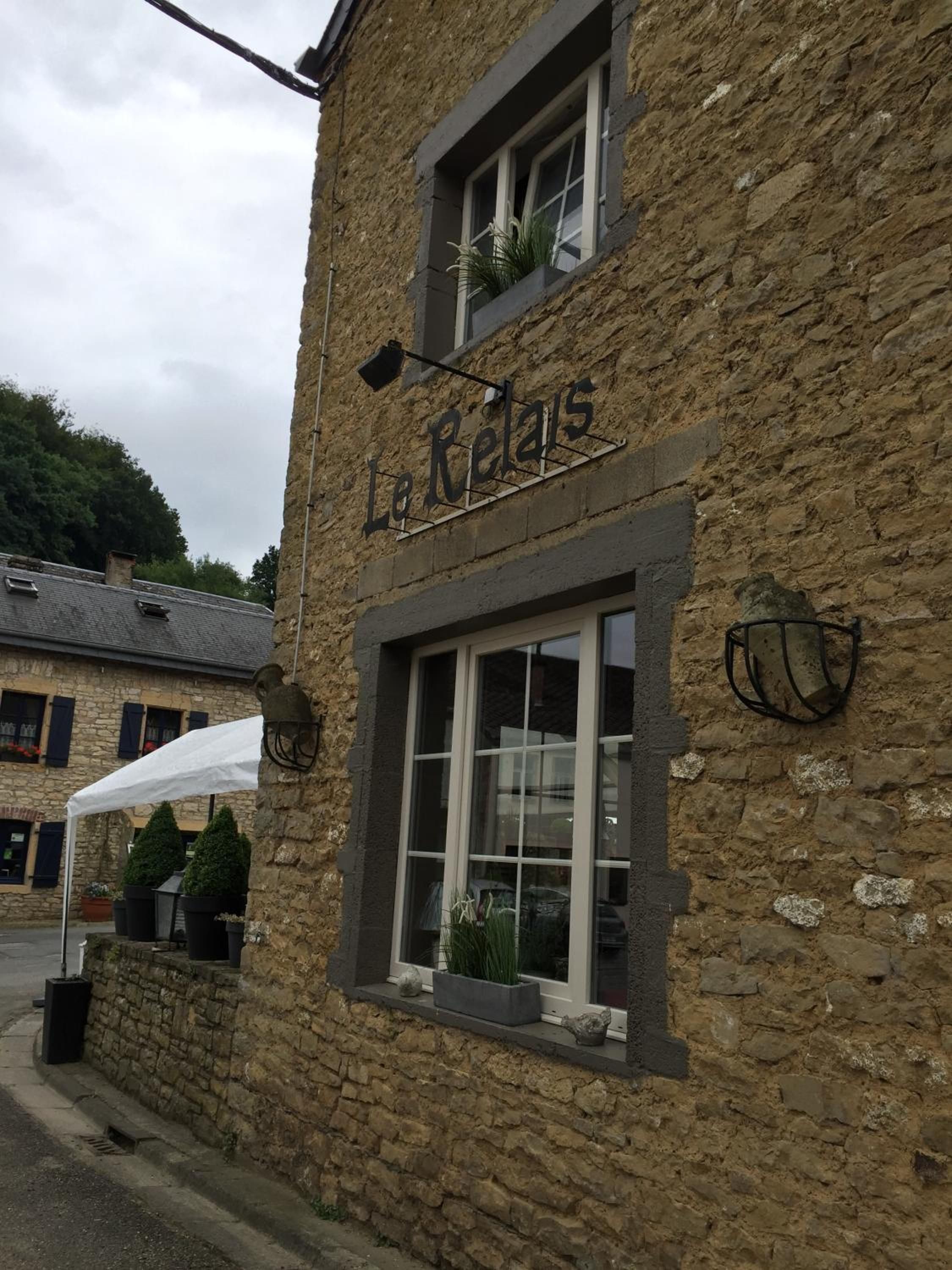 Un bâtiment en pierre avec une enseigne indiquant « Le Relais ». On y voit des fenêtres ornées de plantes, un auvent blanc et un mur en pierre.