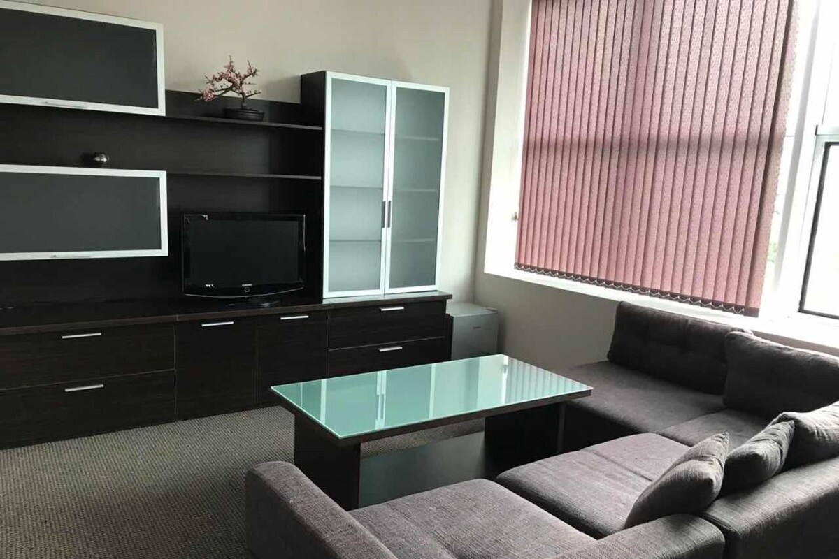 A modern living room with a glass coffee table, grey sofa, dark cabinets, and a television.