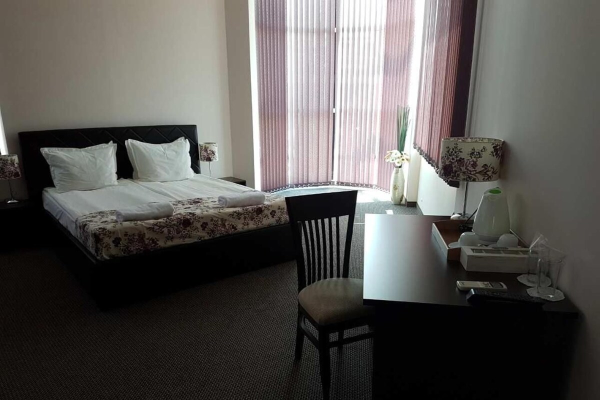 A hotel room with a bed, a desk, a chair, a lamp, and a window with curtains.