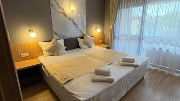 Cama doble con cabecera, dos mesitas de noche y un amplio ventanal con cortinas translúcidas.