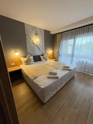 A double bed with a headboard, two bedside tables, and a large window with sheer curtains.