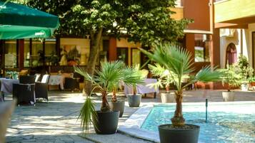 Un área de estar al aire libre con palmeras en macetas, piscina y mesas con sombrillas.