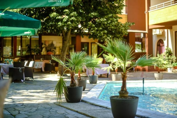An outdoor seating area with potted palm plants, a pool, and tables with umbrellas.