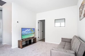 A living room with a flat-screen TV on a wooden stand, a grey sofa, and a door leading to another room. - Tiger Country Retreat in Baton Rouge - CB1008 (Baton Rouge)
