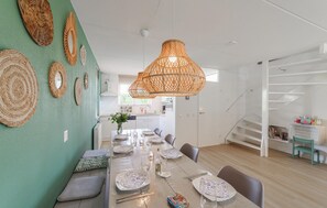 Ein Essbereich mit einem langen Tisch, gedeckt für vier Personen, einer Küche im Hintergrund und einer Treppe, die zu einer weiteren Ebene führt.