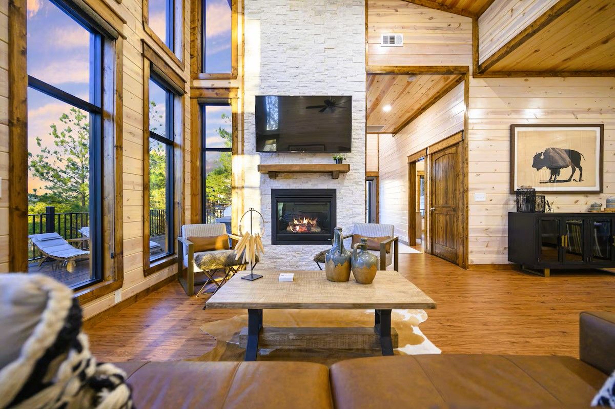 A spacious living room with a fireplace, large windows, and a television mounted above it.
