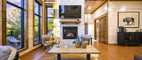 A spacious living room with a fireplace, large windows, and a television mounted above it.