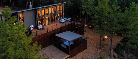 A modern cabin with a deck, a hot tub, and a fire pit surrounded by trees.