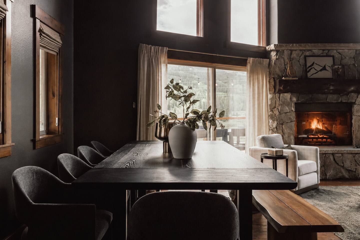 Een eethoek met een lange tafel, stoelen, een bank, een vaas met planten, een open haard en een raam met gordijnen.