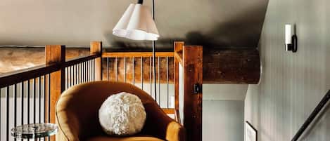 Um quarto acolhedor no estilo loft, com vigas de madeira aparentes, uma poltrona de couro com almofada macia, um banquinho redondo pequeno e uma luminária pendente branca.