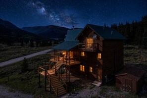 A log cabin with a balcony and a staircase leading to the upper level, surrounded by a mountainous landscape.