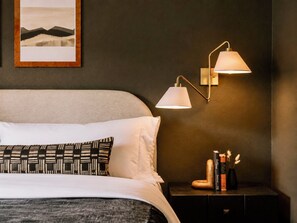 A neatly made bed with white and patterned pillows, a dark wall with a framed picture, and a bedside table with books and a vase of dried flowers.