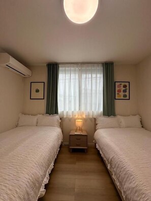 A room with two beds, a bedside table with a lamp, a wall-mounted air conditioner, and a window with curtains. - AND HOME (Osaka)