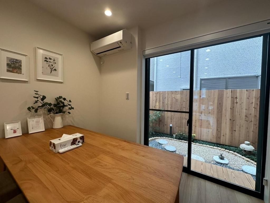 Een kamer met een houten tafel, een airconditioning, ingelijste foto's en uitzicht op een houten schutting en tuin door een schuifdeur.