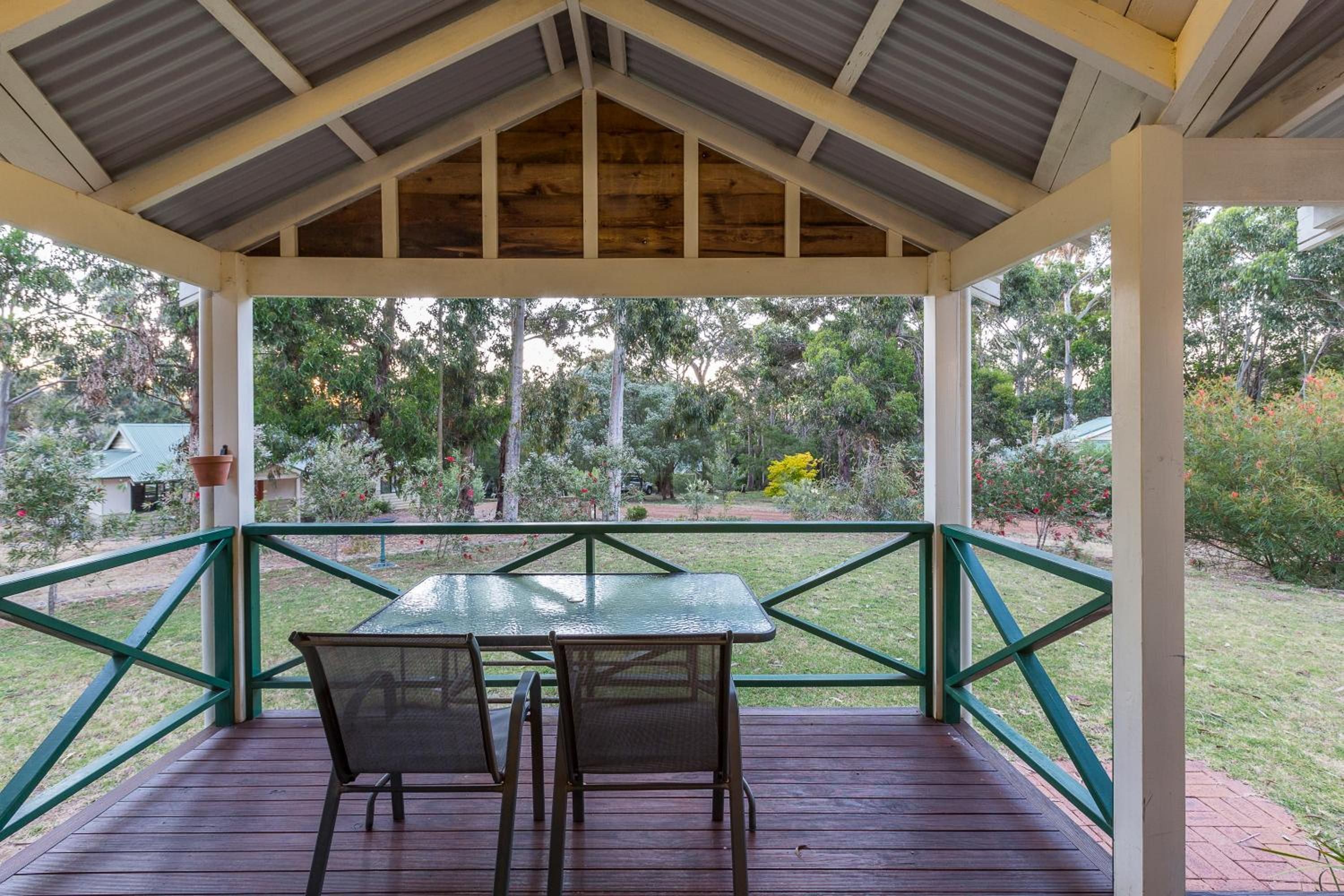 A covered outdoor seating area with a table and chairs, surrounded by a railing and a view of a garden.