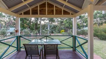 A covered outdoor seating area with a table and chairs, surrounded by a railing and a view of a garden.