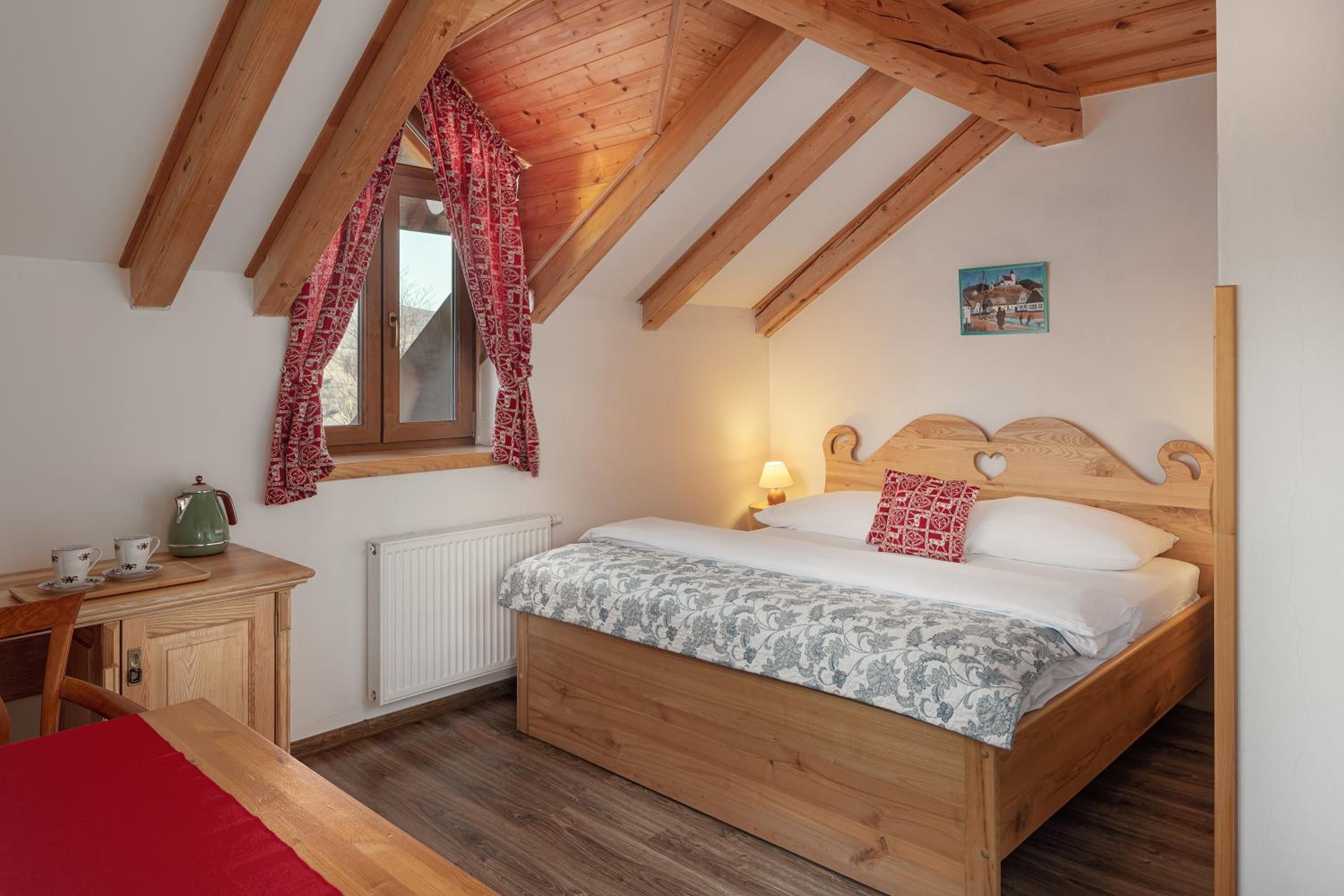 Un dormitorio con cama de madera, un radiador, una ventana con cortinas estampadas, una mesita de noche con tetera y un cuadro en la pared.