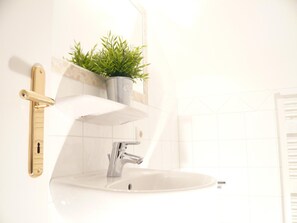 Ein Badezimmer mit einem weißen Waschbecken, einem modernen Wasserhahn und einer Topfpflanze auf einem Regal.