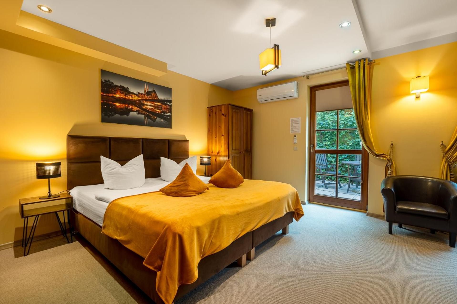 A hotel room with a bed, a nightstand, a chair, an air conditioning unit, and a window with curtains.