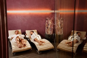 Ein Zimmer mit Spiegel, drei Betten, einer großen Vase mit Trockenpflanzen und einer warmen Lichtleiste an der Wand.