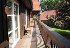Une terrasse en bois avec une rambarde, une chaise et une vue sur un bâtiment dont le toit est recouvert de tuiles rouges.