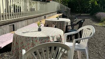 Ett utomhusområde med sittplatser, bestående av runda bord och vita stolar, ett vitt staket och en byggnad med ett rött tak i bakgrunden.