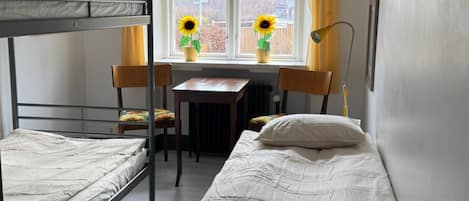 Quarto de dormitório com beliches, escrivaninha, cadeiras, mesa e janela com cortinas.