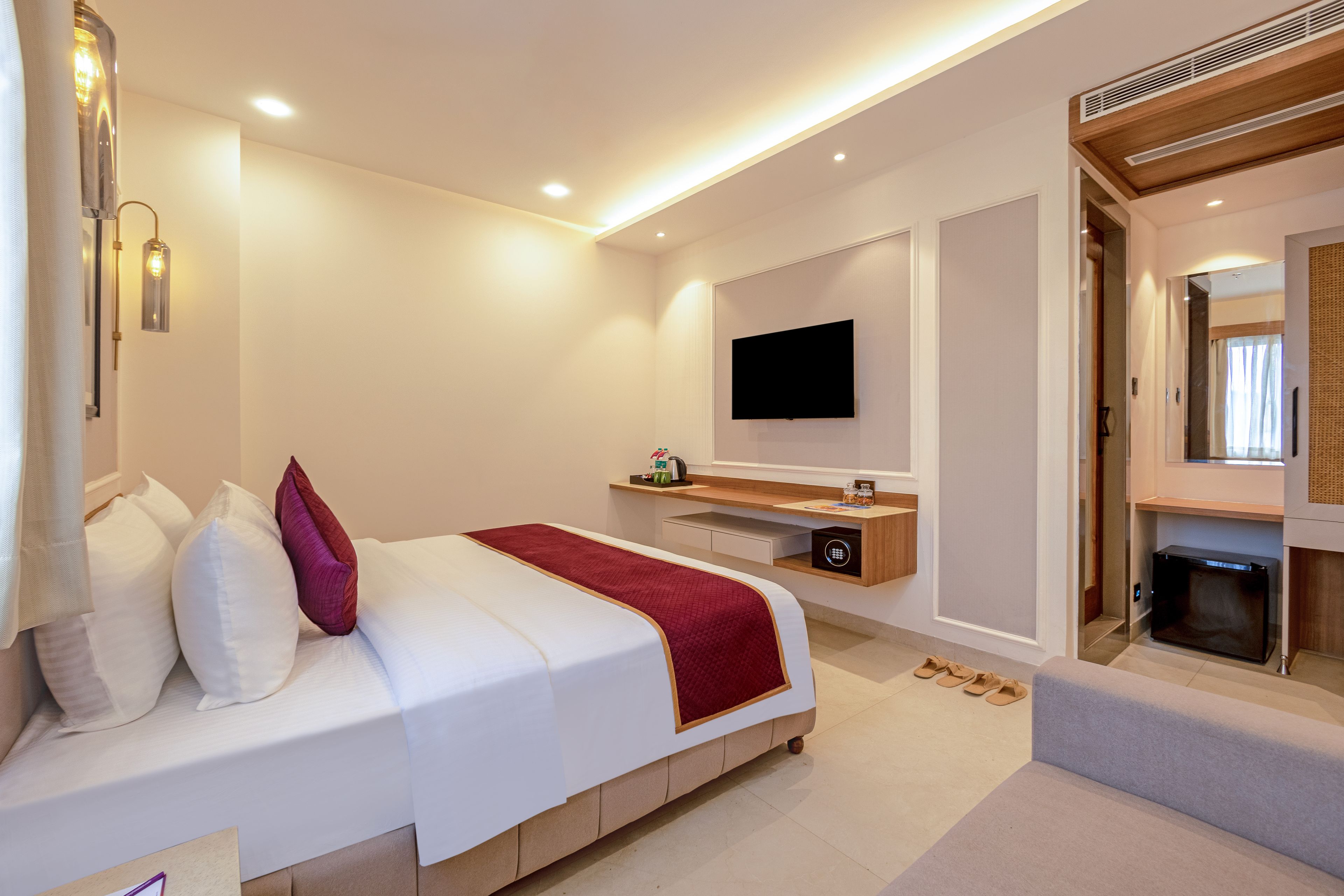 A modern hotel room with a large bed, a flat-screen TV mounted on the wall, a small wooden side table, and a built-in safe.