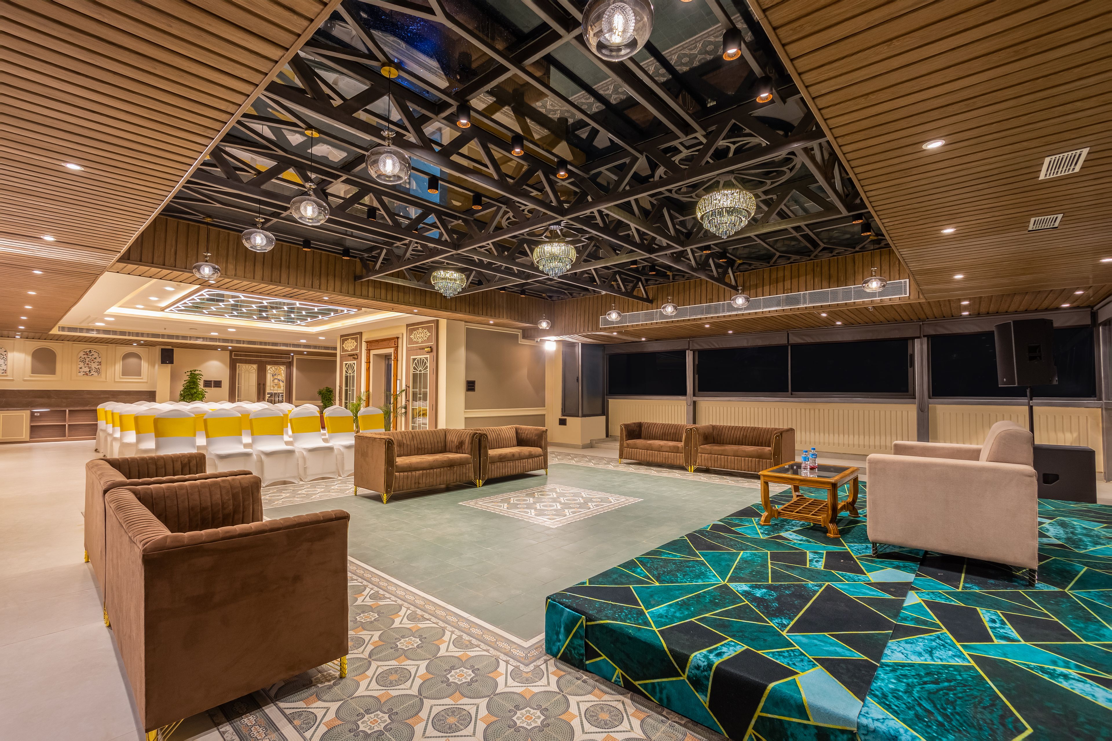 A spacious lounge area with seating arrangements, a patterned carpet, and a high ceiling with exposed beams and lighting fixtures.
