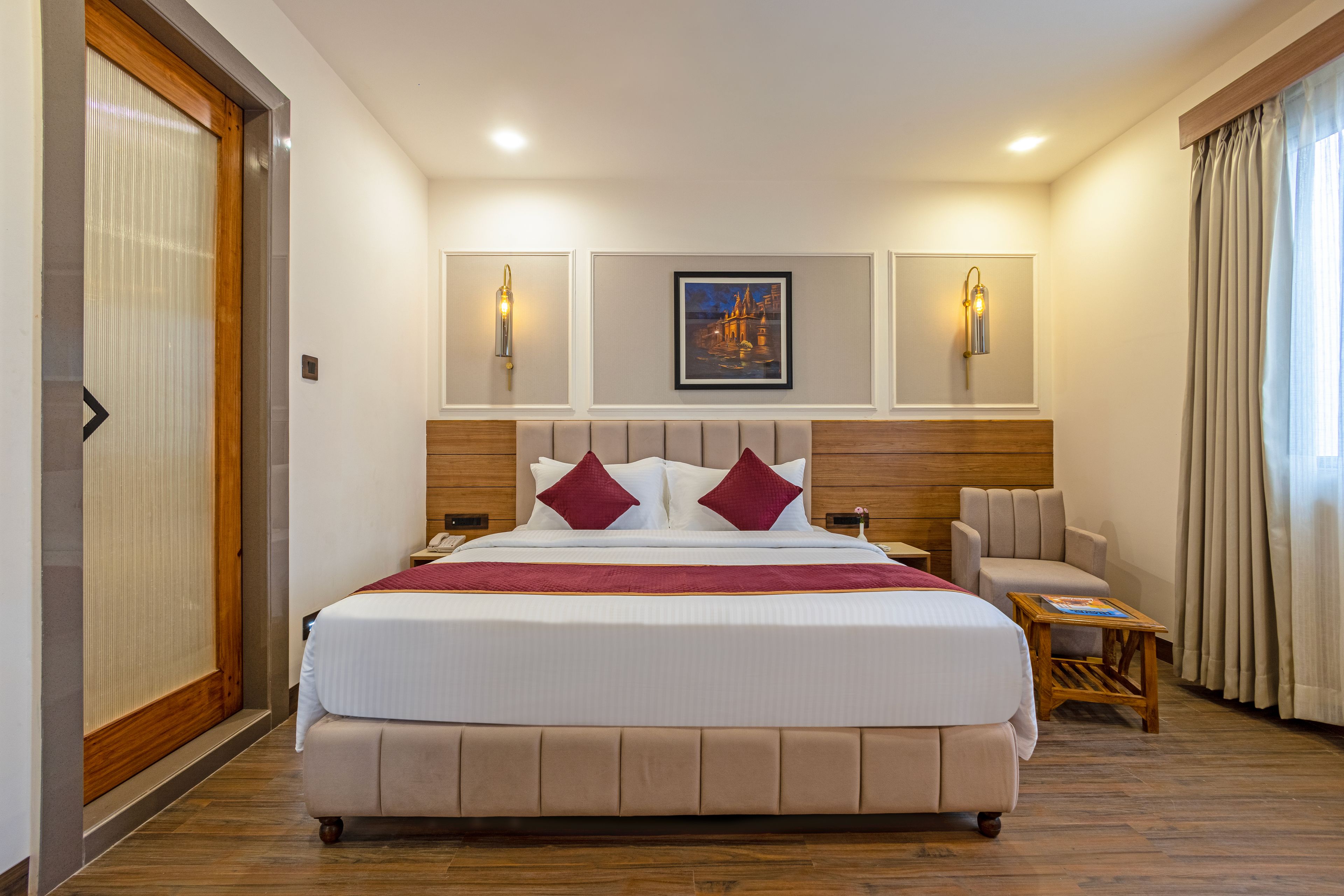 Habitación de hotel con una cama grande, cabecero de madera, dos luces fijadas a la pared, un cuadro enmarco y un rincón de descanso con una silla y una mesita.