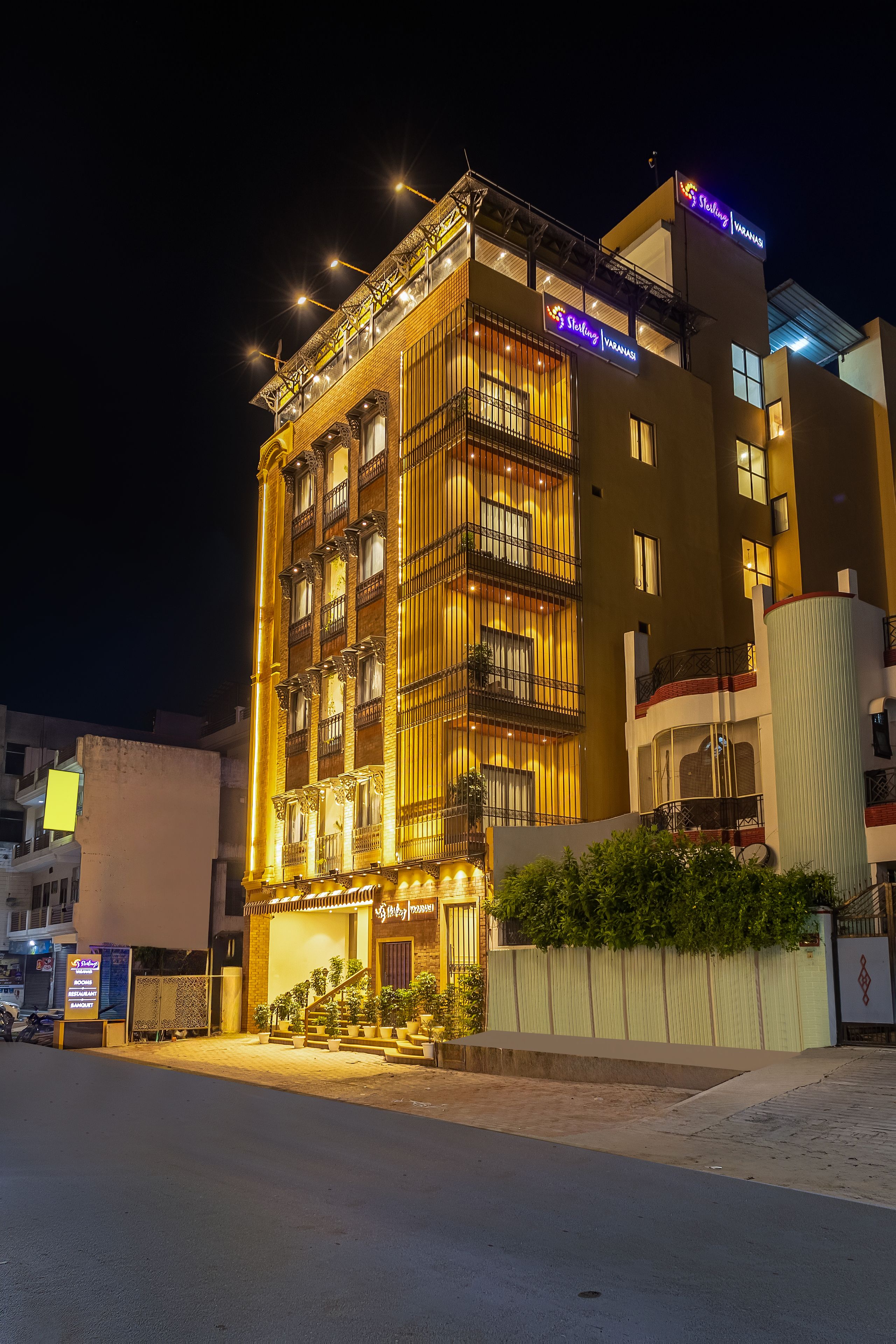 A multi-story hotel with a balcony on the second floor, a sign indicating it is a Sterling Varanasi hotel, and a gated entrance with potted plants.