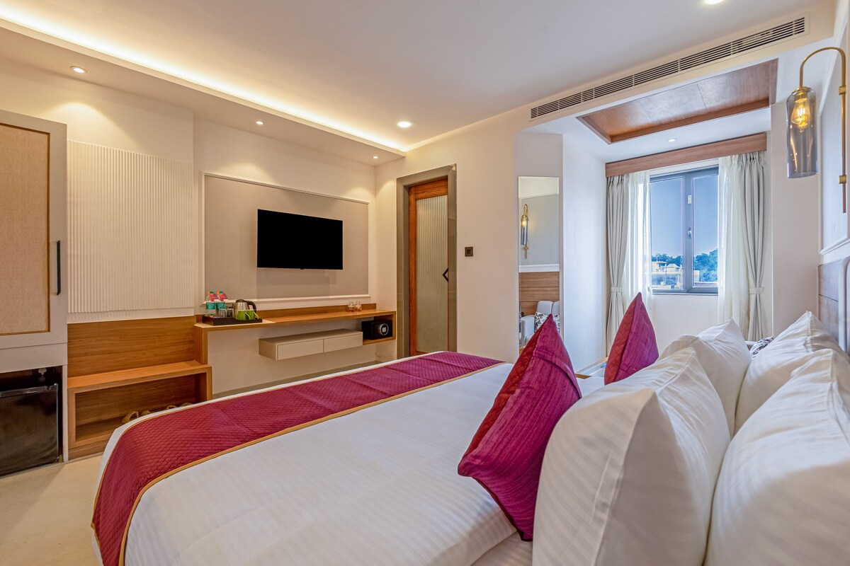Habitación de hotel con una cama grande, un televisor de pantalla plana montado en la pared, una mesita de noche y una ventana con cortinas.