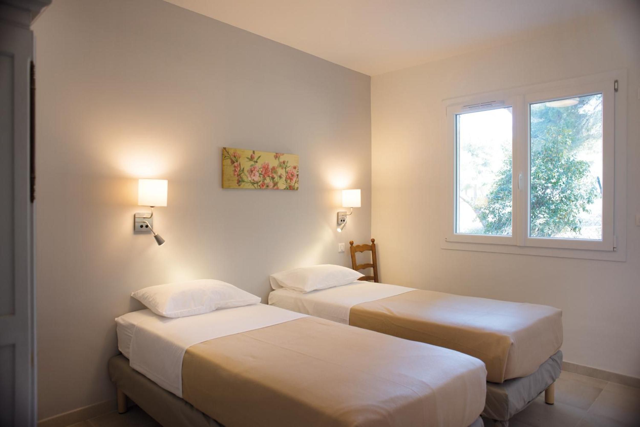A room with two single beds, a wooden chair, a painting on the wall, and a window with a view of trees.