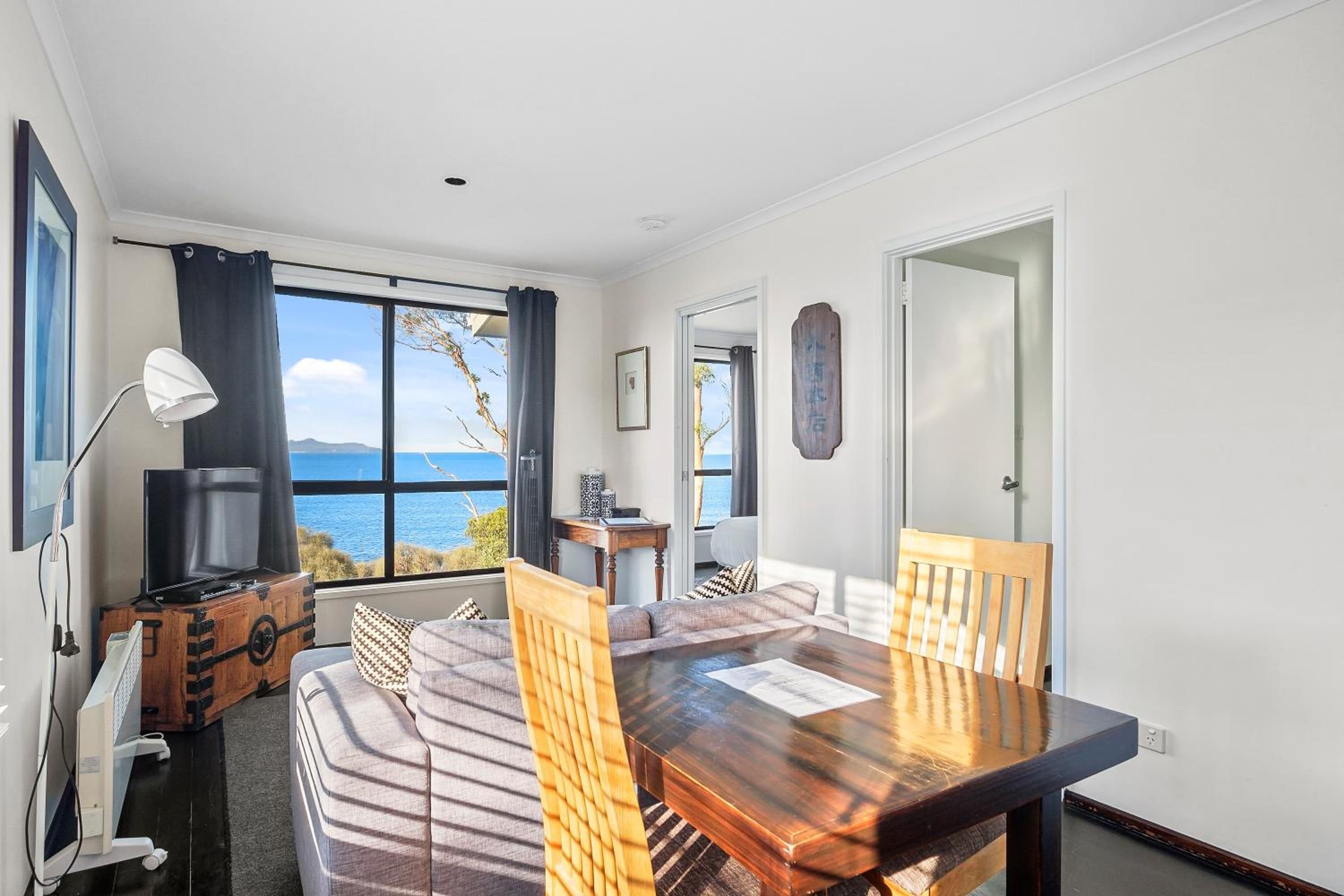 A room with a wooden dining table, two chairs, a sofa, a TV on a wooden cabinet, and a view of the sea through large windows.