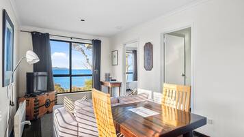 A room with a wooden dining table, two chairs, a sofa, a TV on a wooden cabinet, and a view of the sea through large windows.