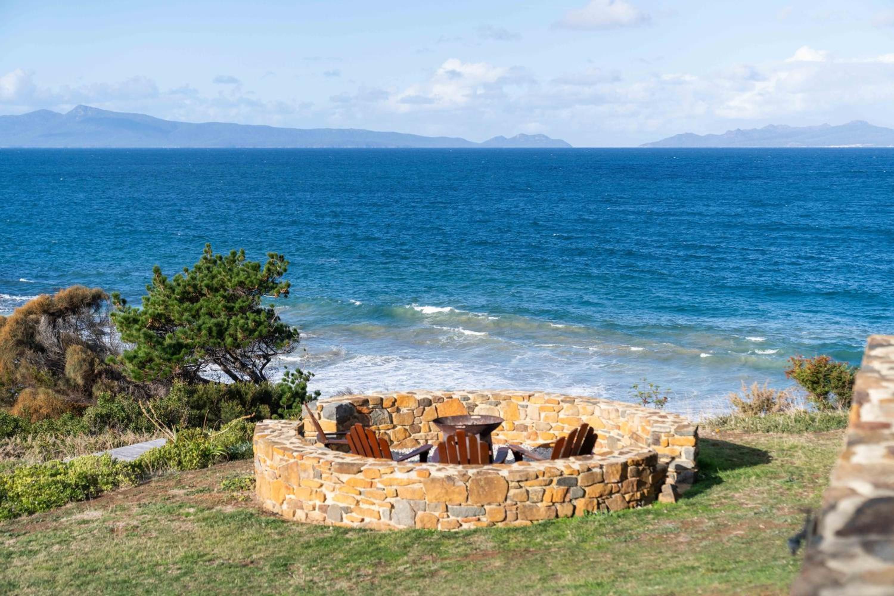 A stone fire pit with seating, overlooking a coastal view with the sea and mountains in the distance.