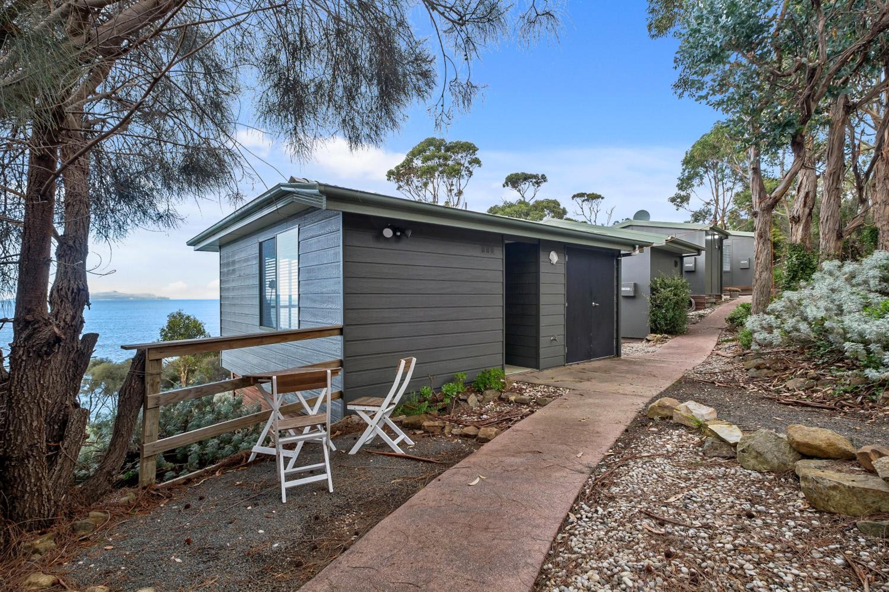 Une cabane moderne dotée d’une terrasse donnant sur un plan d’eau, et entourée d’arbres.