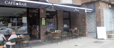 An outdoor café with tables and chairs, a covered entrance, and a sign for 'NIRAS SUPER BOKK'.