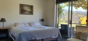 A hotel room with a bed, two bedside lamps, a painting on the wall, and a balcony with a view of the countryside. - Golfview Lodge (Bowral)
