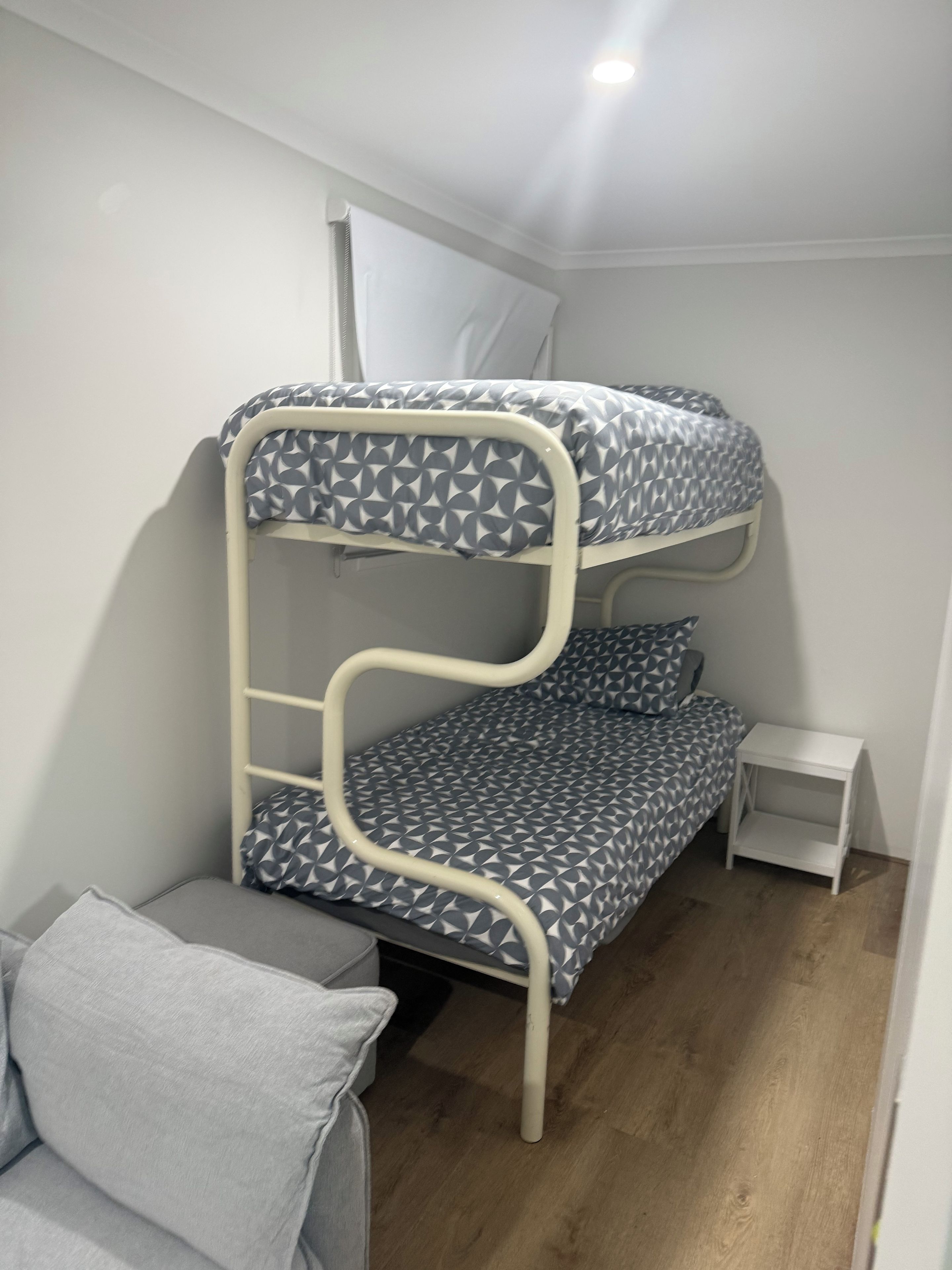 Ein Etagenbett mit einem geometrisch gemusterten Bettbezug, ein kleiner Nachttisch und ein graues Sofa in einem Zimmer mit Holzboden und weißen Wänden.