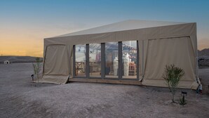 Una tenda gran de color beix amb una porta de vidre i finestres, situada en un desert amb vegetació escassa i un cel serè.