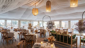 A tented dining area with tables set for a meal, wooden chairs, and hanging light fixtures.