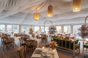 A tented dining area with tables set for a meal, wooden chairs, and hanging light fixtures.