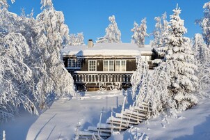 Una cabaña de madera cubierta de nieve con una escalera visible que lleva a la entrada, rodeada de árboles cargados de nieve.