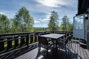 Una terraza de madera con una mesa y sillas, con vista a un paisaje con árboles y una lejana perspectiva.