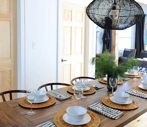 Een eettafel gedekt met borden, kommen en bestek, een vaas met groen, en een zwarte lamp met een net.