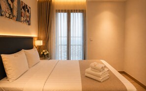 A hotel room with a large bed, a nightstand with a lamp, a vase with flowers, and a view of the city through the window. - Zenity Luxury Central HCM by BEARHOUSE (phuong cau ong lanh)