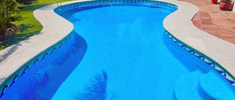 A swimming pool with a tiled edge, surrounded by greenery and a building with a tiled roof.
